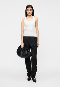 Γυναίκα με λευκό τοπ χωρίς μανίκια και μαύρο παντελόνι, κρατώντας μια μεγάλη μαύρη δερμάτινη τσάντα χειρός, στέκεται μπροστά σε απλό λευκό φόντο.