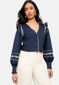 Navy blouse with ruffled shoulders, floral embroidery, and scalloped edges. Features buttons and fitted cuffs with striped detail.