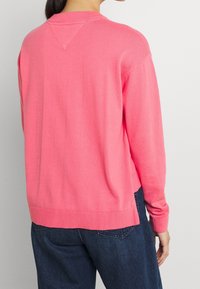 Korallrosa tröja, stickad textur, ribbad linning och nederkant, avslappnad passform och lätt längre bakre nederkant. Buren över mörkblå jeans.