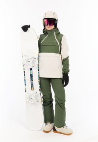 Persoon in een groen en beige snowboardoutfit die een witte snowboard met kleurrijke strepen en witte bindingen vasthoudt.
