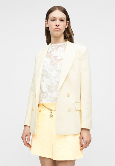 Femme portant un blazer croisé crème sur un chemisier en dentelle florale blanche et une jupe jaune texturée avec une ceinture à chaîne dorée.