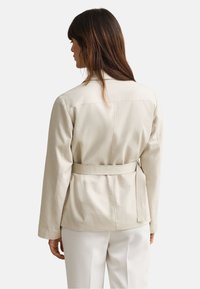 Γυναίκα με καστανά μαλλιά που φοράει μπεζ σακάκι με ζώνη και λευκό παντελόνι, εμφανίζεται από πίσω με φόντο έναν απλό φωτεινό τοίχο.