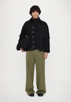 Black oversized shirt with white stitching and buttons, paired with wide olive green trousers and black shoes. Holding a small black bag.