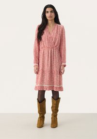 Vestido floral con escote en V, mangas largas y un dobladillo fluido, con patrones rojos y blancos. Combinado con botas de ante marrón hasta la rodilla.