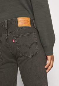 Jeans de ganga cinza escuro com um corte reto clássico. Apresenta um retalho de pele castanha e uma etiqueta vermelha no bolso traseiro. Tecido texturizado.