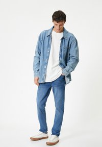 Camisa de mezclilla azul claro sobre una camiseta blanca, combinada con jeans azul claro y zapatillas blancas con suelas de goma marrón. Diseño sencillo, ajuste relajado.