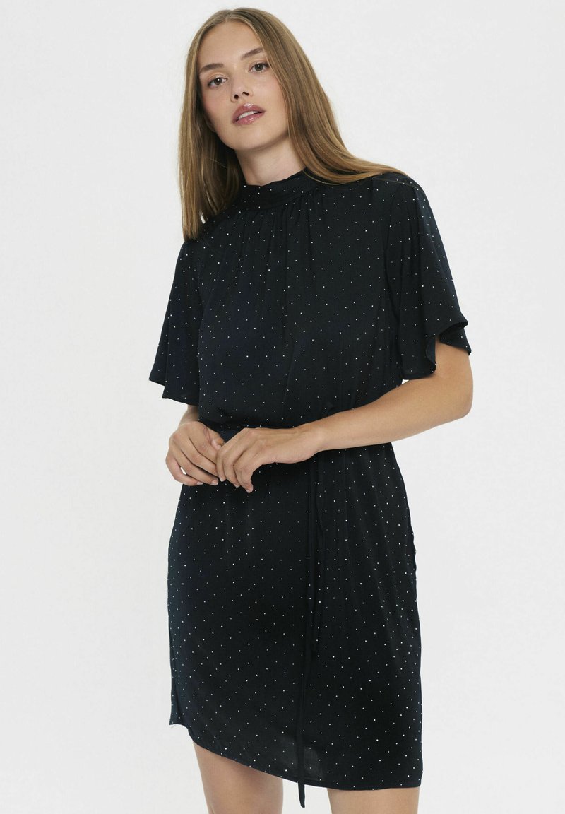 Vestido corto negro con un escote elástico alto, mangas de volantes y un patrón de lunares blancos. Presenta un detalle de cintura atada y una tela suave.
