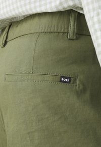 Pantalon vert olive en tissu lisse, avec une poche arrière ornée d'une étiquette noire "BOSS" et des détails de couture subtils.