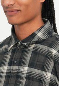 Camicia a quadri bianco e nero con colletto a bottoni, realizzata in tessuto testurizzato, caratterizzata da ampi motivi a scacchi e due bottoni visibili sulla patta.