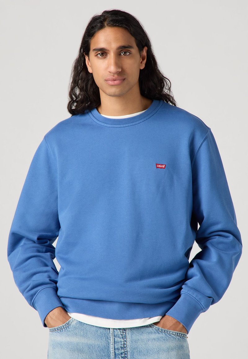 Sweatshirt bleu à col ras du cou en tissu doux, arborant un petit logo rouge Levi's sur la poitrine et des poignets côtelés. Porté sur une chemise blanche.