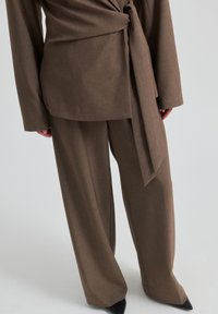 Pantalon de costume marron sur mesure associé à une veste assortie avec un long nœud latéral, porté avec des chaussures noires à bout pointu sur un fond blanc.
