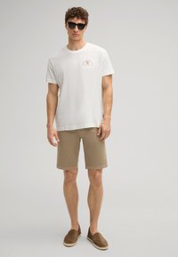 Camiseta blanca texturizada con pequeño logotipo marrón, pantalones cortos beiges y espadrilles marrones. El modelo también lleva gafas de sol negras.