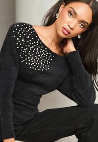 Femeie îmbrăcată cu un pulover negru pufos, cu studs decorative argintii grupate aproape de umăr, pantaloni negri și cercei argintii, așezată cu o mână aproape de gât.