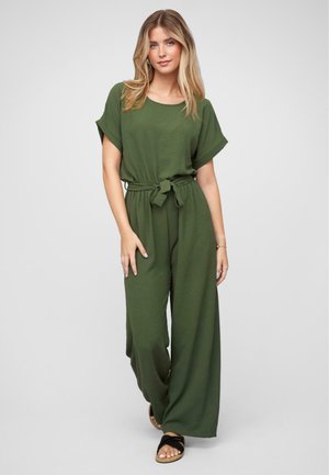 Donna con lunghi capelli biondi che indossa una tuta verde oliva ampia con maniche corte, una cintura legata in vita e sandali neri con punta aperta.