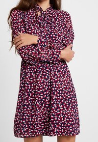 Blommig klänning med en marinblå bas och ett mönster av röda och vita tårformade former. Långa ärmar, knytkrage och avslappnad passform.