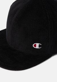 Schwarze Cordmütze mit flachem Schirm. Mit gesticktem Logo in Rot und Weiß an der Seite. Textil mit vertikalen Streifen.