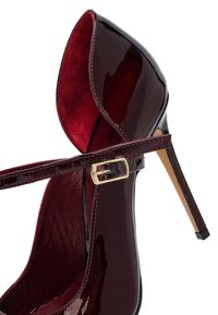 Zapato de tacón alto de charol rojo oscuro con punta afilada y tirante delgado en el tobillo que presenta una hebilla dorada, mostrado sobre un fondo blanco.