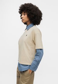 Μπεζ κοντομάνικη μπλούζα φορεμένη πάνω από μπλε ριγέ μακρυμάνικο πουκάμισο, συνδυασμένη με χακί παντελόνι. Διαθέτει μικρό λογότυπο στην μπλούζα.