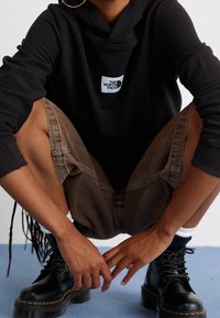 Felpa nera con cappuccio, texture a coste e patch con logo. Shorts di jeans marroni con dettagli sui taschini, abbinati a stivali neri con lacci.