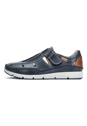 Zapato de cuero azul oscuro con cortes laterales, suela moteada blanca y adornos de cuero marrón. Presenta una correa segura en la parte superior.