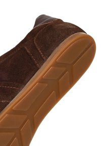 Suola di scarpa in camoscio marrone scuro inclinata verso l'alto, che mostra la suola esterna in gomma marrone chiaro con motivi geometrici di battistrada.