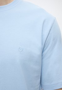 Φωτεινό γαλάζιο μπλουζάκι με κοντά μανίκια και στρογγυλή λαιμόκοψη, με ένα μικρό κεντητό λογότυπο κύκλου και κάθετης γραμμής στο αριστερό στήθος.