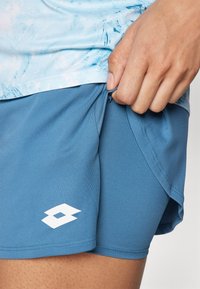 Gaiši zils sporta krekls ar abstraktu rakstu, apvienots ar zilām šortiem ar baltu logo. Audums izskatās vienmērīgs un viegls.