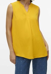 Ärmellose gelbe Bluse mit V-Ausschnitt, die eine strukturierte Oberfläche aufweist. Das Design ist locker geschnitten und wird mit verzierten Jeans kombiniert.