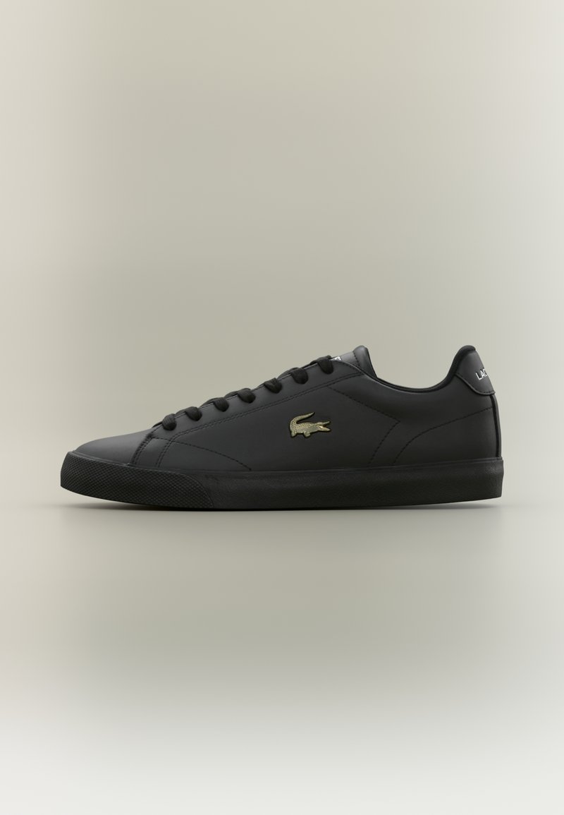 Sorte lavtop sneakers laget av glatt syntetisk materiale, med gummisåle, flate lisser og en gull logo detalj på siden.