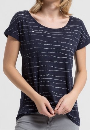 Femme portant une chemise bleu marine à manches courtes avec des lignes ondulées blanches et de petits motifs de bateaux en papier, tenant l'ourlet de la chemise.