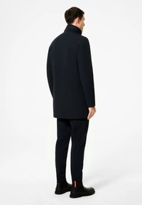 Cappotto blu navy con colletto alto, design elegante e texture liscia, abbinato a pantaloni scuri e stivali neri con un accento arancione.