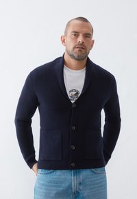 Navy ribbed cardigan with a shawl collar, front buttons, and two patch pockets, worn over a white T-shirt with embroidered detail.