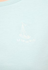 Světle modré tričko s kulatým výstřihem, které má bílé vyšívané design plachetnice a text "LA VIE EN BLEU" níže. Měkká textura.