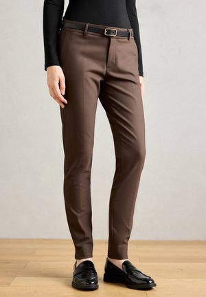 Henkilö, jolla on ylläsäetut ruskeat housut, musta vyö, musta pitkähihaista paita ja kiiltävät mustat loafersit, seisoo puulattialla.