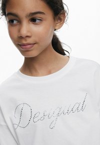 Weißes Baumwoll-T-Shirt mit dem Schriftzug "Desigual", verziert mit kleinen Strasssteinen in einer geschwungenen Schriftart. Rundhalsausschnitt, kurze Ärmel.
