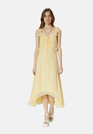 Jonge vrouw die een mouwloze, lichtgele high-low jurk met ruches en witte platformsandalen draagt, staand tegen een effen achtergrond.