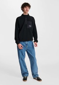 Sort fleecepullover med kvartlynlås ved halsen, frontlomme med en kontrastlogo, kombineret med lyseblå baggy jeans og sorte sneakers.