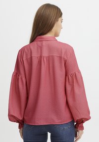 Rote gemusterte Bluse mit Kragen, ballonförmigen langen Ärmel und gepuffertem Rückendetail. Hergestellt aus leichtem Stoff.