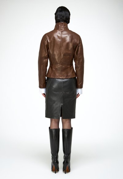 Kvinde iført en tætsiddende brun læderjakke, sort lædernederdel med slids bagpå, gennemsigtige strømpebukser og sorte knæhøje støvler, stående med ryggen til.
