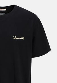 Camiseta de algodón negra con escote redondo, mangas cortas y bordado frontal de "Originals" en hilo dorado. Textura suave.