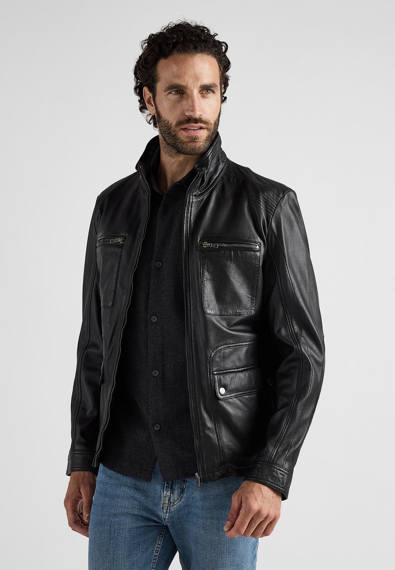 Schwarze Lederjacke mit Stehkragen, zwei Reißverschlusstaschen auf der Brust und einer strukturierten linken Tasche, kombiniert mit einem dunklen Hemd mit Knöpfen.
