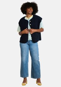 Gilet in pile blu con bottoni grandi, indossato sopra una camicia a righe chiara. Abbinato a jeans blu e scarpe gialle con tacco a blocco.