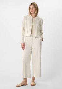 Jasnobeżowa krótka kurtka zapinana na zamek, zestawiona z szerokimi beżowymi spodniami i plecionymi klapkami, na jednolitym tle.