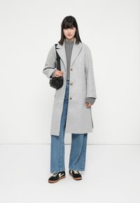 Donna che indossa un lungo cappotto grigio chiaro, un maglione grigio con zip, jeans a gamba larga blu, scarpe da ginnastica nere e porta una piccola borsa a spalla nera.