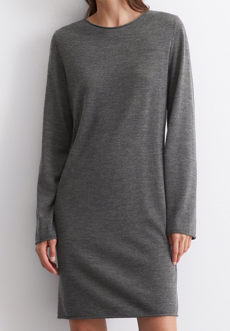 Femme portant une robe en maille grise unie, manches longues, longueur genou, avec un col rond, debout sur fond blanc.