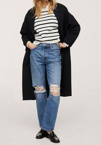 Jean bleu déchiré, pull rayé noir et blanc, et un long manteau noir. Tissu lisse, coupe décontractée, style casual. Des chaussures noires complètent le look.