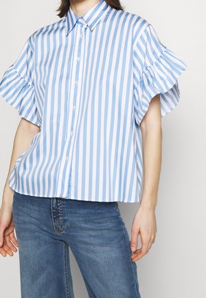 Personne portant un chemisier à manches courtes rayé verticalement bleu et blanc avec des détails à volants, associé à un jean bleu en denim.