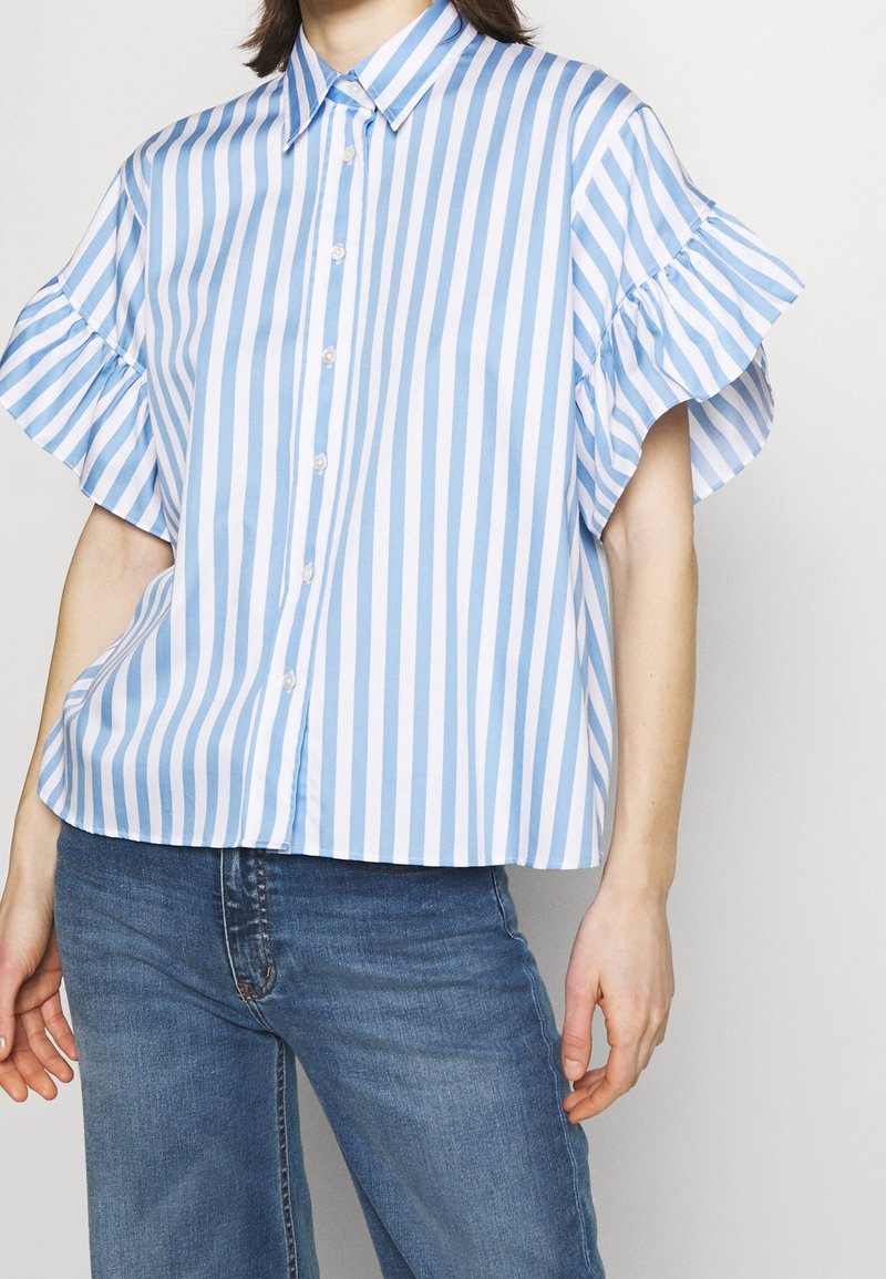 Personne portant un chemisier à manches courtes rayé verticalement bleu et blanc avec des détails à volants, associé à un jean bleu en denim.