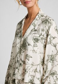 Vrouw in een crèmekleurige blouse met knopen, voorzien van een gedetailleerde zwarte bloemen- en vlinderafdruk, met een borstzak en een brede kraag.