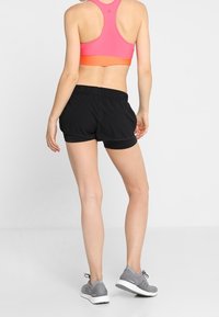 Pantalones cortos deportivos negros con una cinturilla elástica, combinados con un sujetador deportivo rosa. Modelo vistiendo zapatillas de correr grises, mostrando una vista trasera.